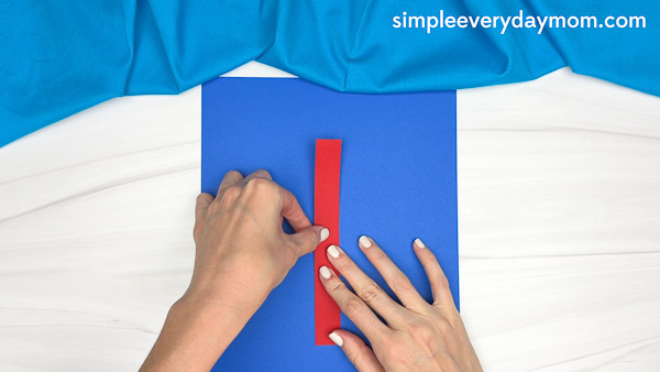 Hands gluing a red paper strip vertically onto a blue sheet for a simple apple core craft for kids.