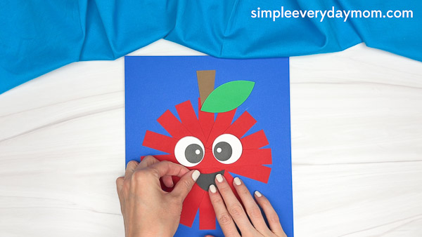 Hands gluing a black paper mouth onto a red paper strip apple with eyes, stem, and leaf on a blue background.