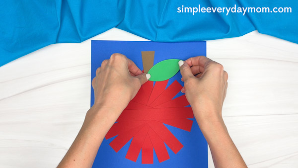 Hands gluing a green paper leaf to the top of a red paper strip apple craft on a blue background.