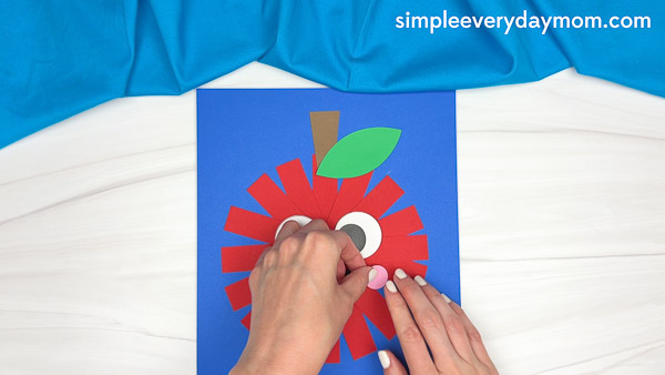 Hands gluing pink paper cheeks onto a red paper strip apple craft with large eyes, stem, and leaf on a blue background.