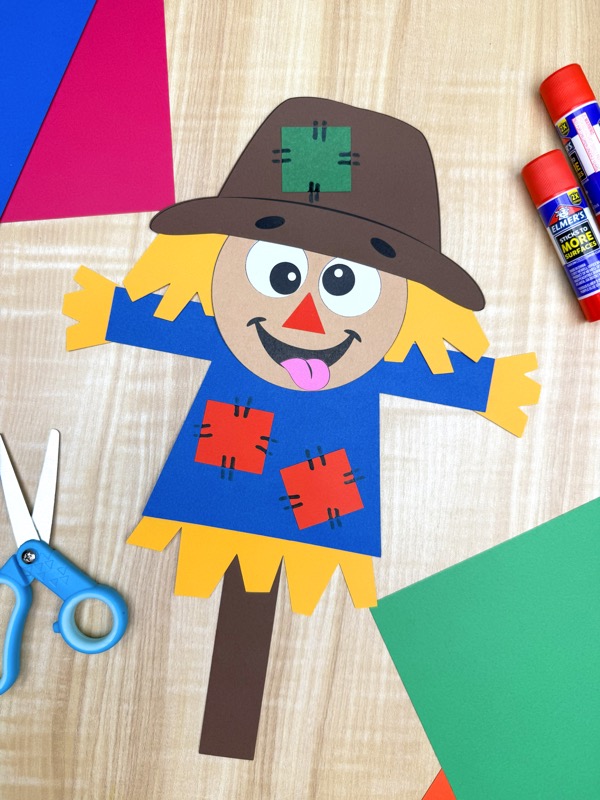 Completed paper scarecrow craft on a table surrounded by scissors, glue sticks, and colored paper. Fun fall activity for kids using printable templates.