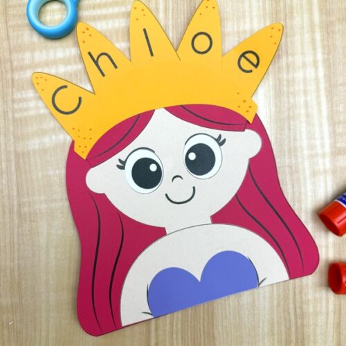 Mermaid name craft featuring a paper mermaid with red hair, purple top, and a yellow crown labeled “Chloe,” displayed on a wooden table with scissors and glue stick nearby – fun and simple name activity for kids.