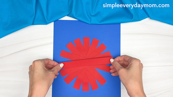 Hands assembling red paper strips into a round shape to create a paper strip apple craft on a blue background.