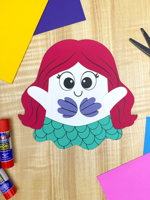 A completed mermaid ghost disguise craft with red hair, a purple seashell bra, and a teal tail. Surrounding the craft are Elmer's glue sticks, scissors, and colorful paper in pink, purple, and yellow.