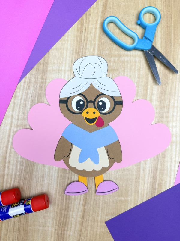 Completed grandma turkey paper craft with glasses, white hair, blue shawl, pink shoes, and large pink feathers, displayed on a wooden surface with scissors, glue sticks, and colored paper.