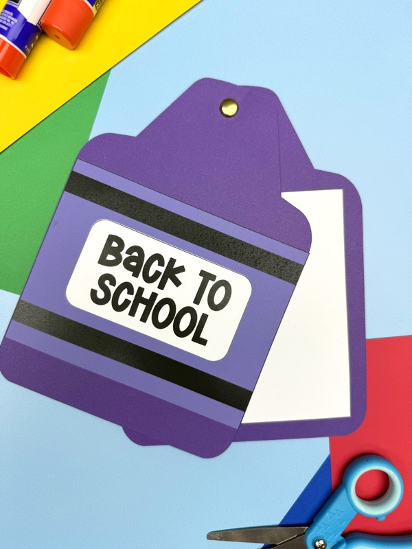 A finished purple crayon book craft with a white blank page that opens from the side, scissors, and glue sticks are on a craft table.
