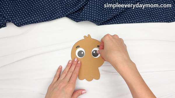 Hands gluing paper googly eyes onto a brown paper turkey head cutout for a Thanksgiving craft.