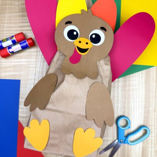 A completed craft of an adorable paper bag turkey with colorful paper tail feathers on a wooden background with craft supplies.