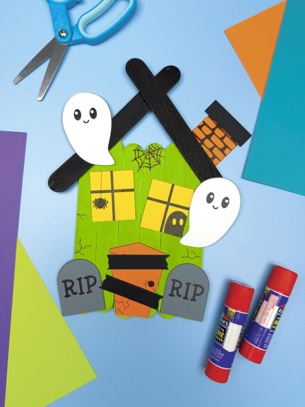A completed craft is a Halloween haunted house made from green popsicle sticks with a black popsicle stick roof, two smiling ghosts, a spider, two gravestones, and a black and orange brick chimney. Craft supplies like scissors, glue sticks, and colored paper are also visible on a blue background.