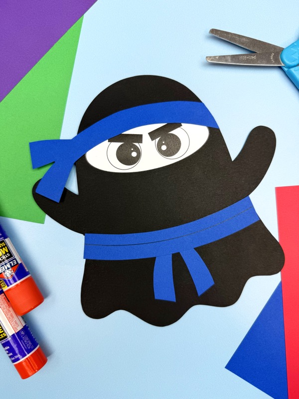 A finished ninja ghost craft with a blue headband and belt sits on a light blue background surrounded by colorful paper and glue sticks.