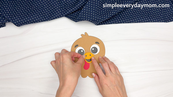 Hands gluing a yellow paper beak and orange mouth to the turkey's face cutout, completing the facial features.