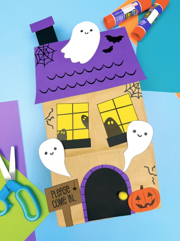 A completed Craft of a Halloween haunted house paper bag with ghosts, a pumpkin, bats, and scissors and glue sticks nearby.