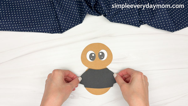 Hands applying a black shirt cutout to a brown paper craft body with attached eyes.