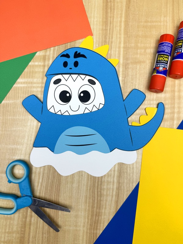A completed craft featuring a cute blue dinosaur ghost cutout with yellow spikes on its back and tail, surrounded by craft paper and supplies on a wooden surface.