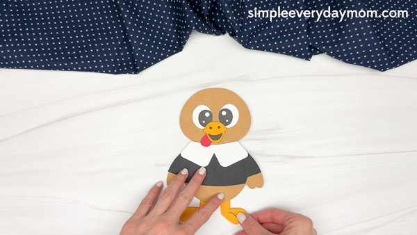 Hands adding orange legs and feet to the bottom of the paper pilgrim turkey craft.