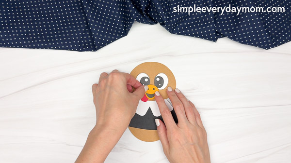 Hands placing a red snood and a yellow beak onto the paper pilgrim's face.