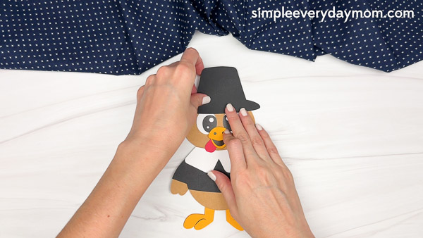 Hands securing a black pilgrim hat to the top of the paper turkey craft's head.