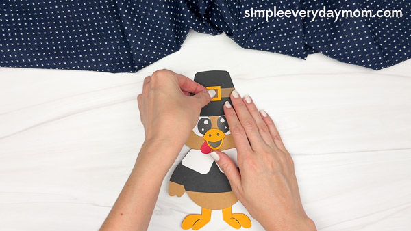 Hands securing a small orange buckle to the band of the black pilgrim hat on the turkey craft.