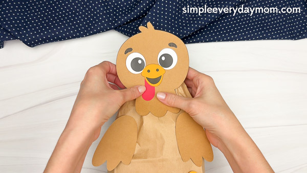 Hands gluing the completed brown paper turkey head onto the paper bag body for a Thanksgiving craft.