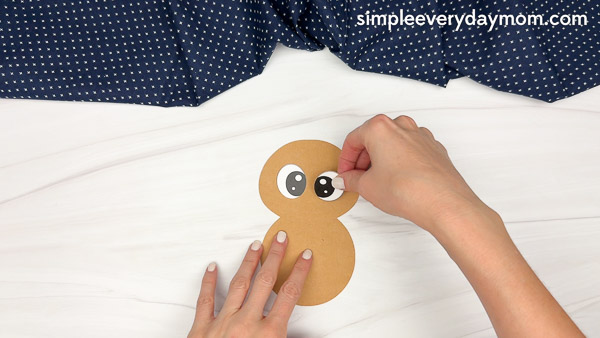 Hands attaching googly eyes to a brown paper craft in the shape of the number eight.