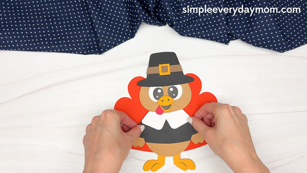 Hands gluing paper feathers behind the paper pilgrim turkey craft's body.