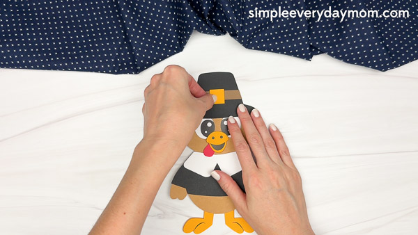 Hands applying a small orange buckle to the band of the black pilgrim hat on the turkey craft.