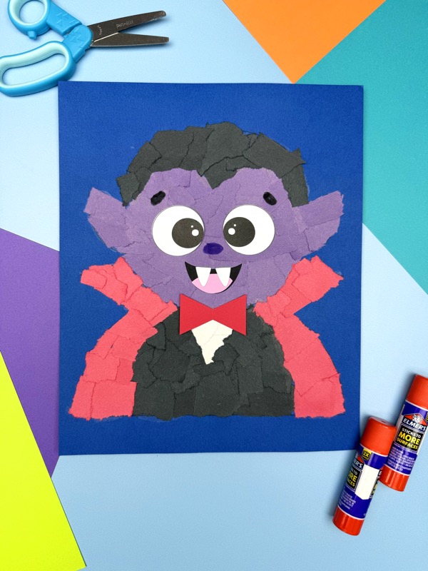 A ripped paper vampire on a blue background, surrounded by scissors, glue sticks, and colorful paper.