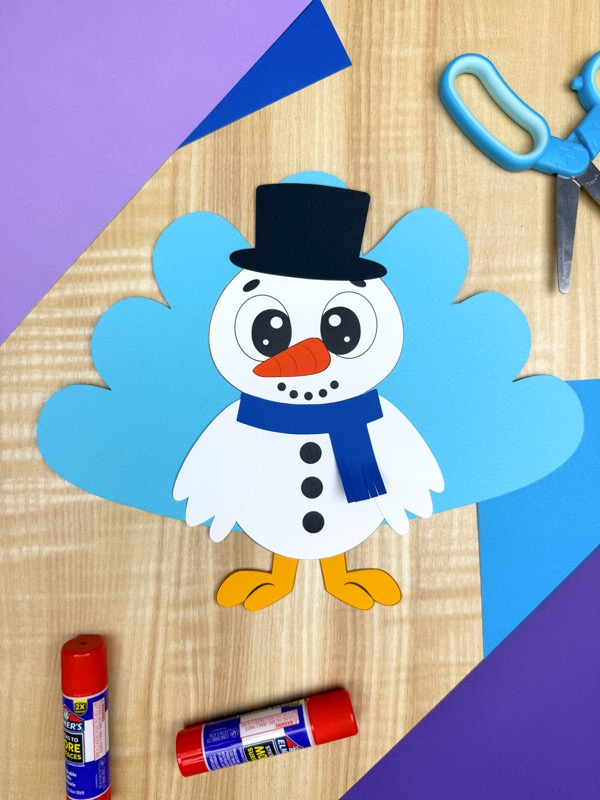 A cheerful snowman turkey disguise paper craft with a black top hat and blue scarf is centered on a light wooden background, surrounded by crafting tools like scissors and glue sticks, and colorful purple and blue construction paper, great for a DIY winter activity.