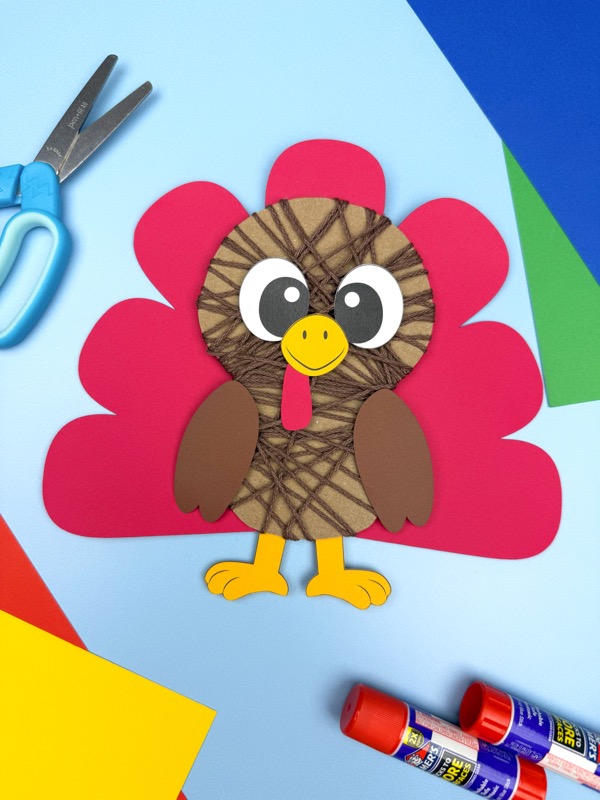 Yarn-wrapped turkey craft on a blue background with magenta tail feathers, surrounded by scissors and glue sticks, an easy fall project.
