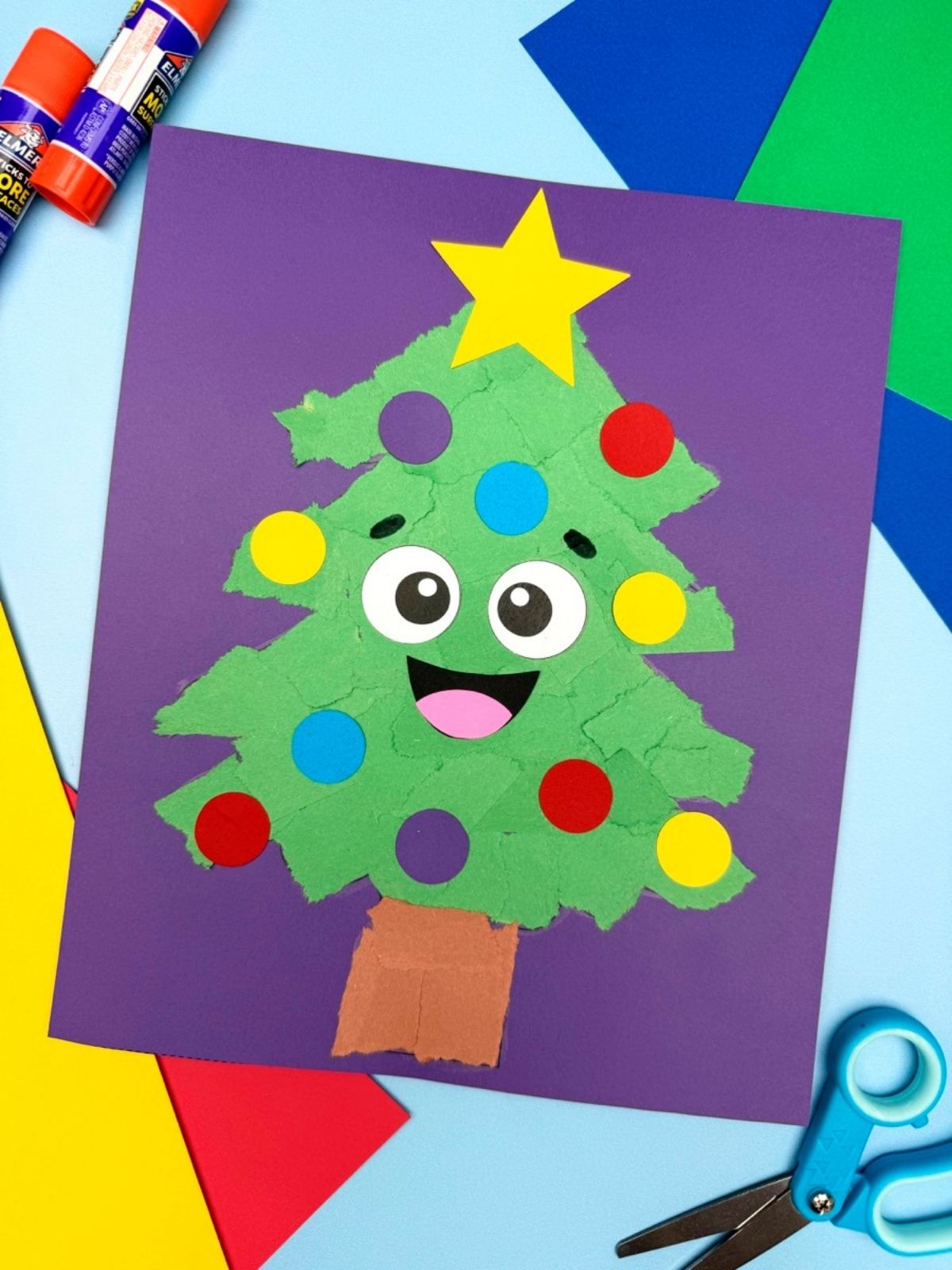 A completed craft of a happy torn paper Christmas tree on a purple background, surrounded by glue sticks, scissors, and colorful construction paper.