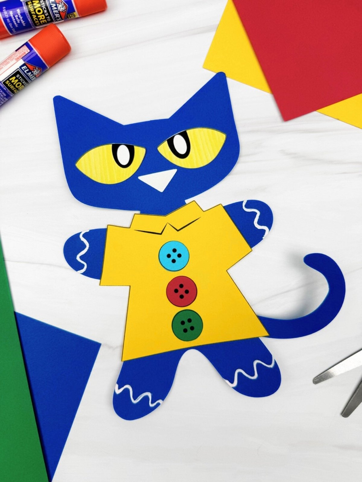 A completed craft of the Pete the Cat gingerbread disguise made from paper, surrounded by craft supplies like blue, green, red, and yellow cardstock, glue sticks, and scissors.
