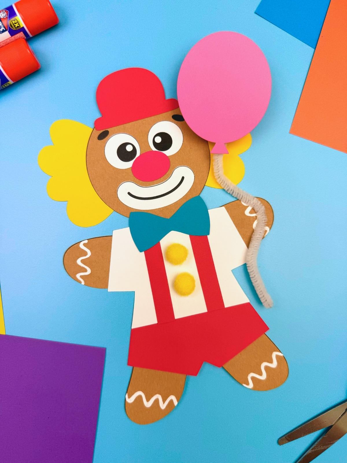 A completed craft showing a clown gingerbread man disguise with red outfit, yellow buttons, blue bow tie, and pink balloon on a blue background for a fun kids Christmas gingerbread craft.