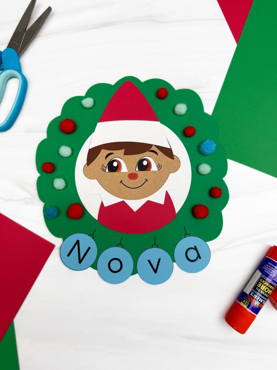 A completed craft showing an Elf on the Shelf name wreath decorated with colorful pom poms and blue letter ornaments spelling Nova for a festive Christmas kids activity.