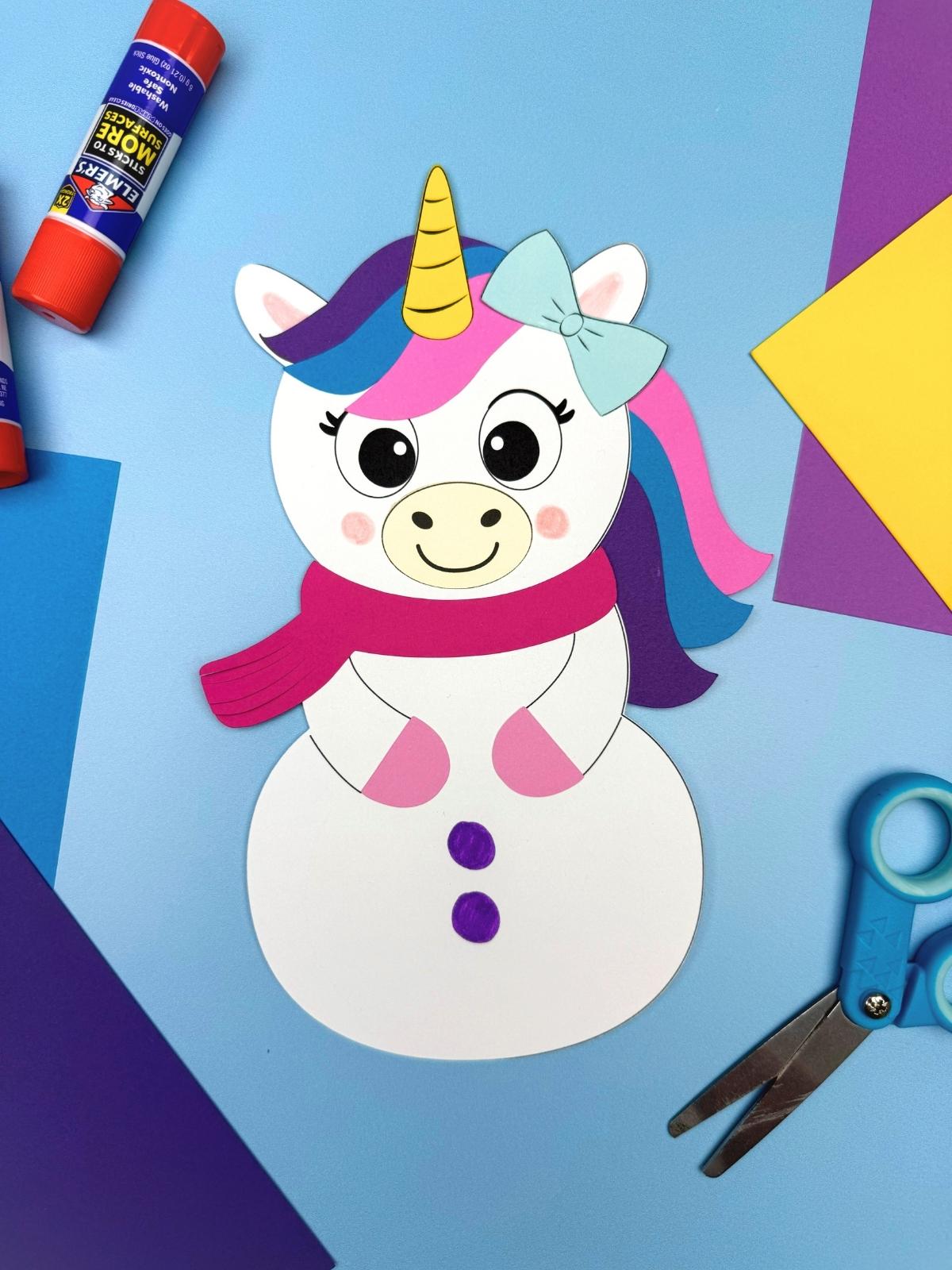 A completed craft showing a unicorn disguise snowman with a colorful mane, horn, scarf, and drawn buttons surrounded by simple paper craft supplies for kids.