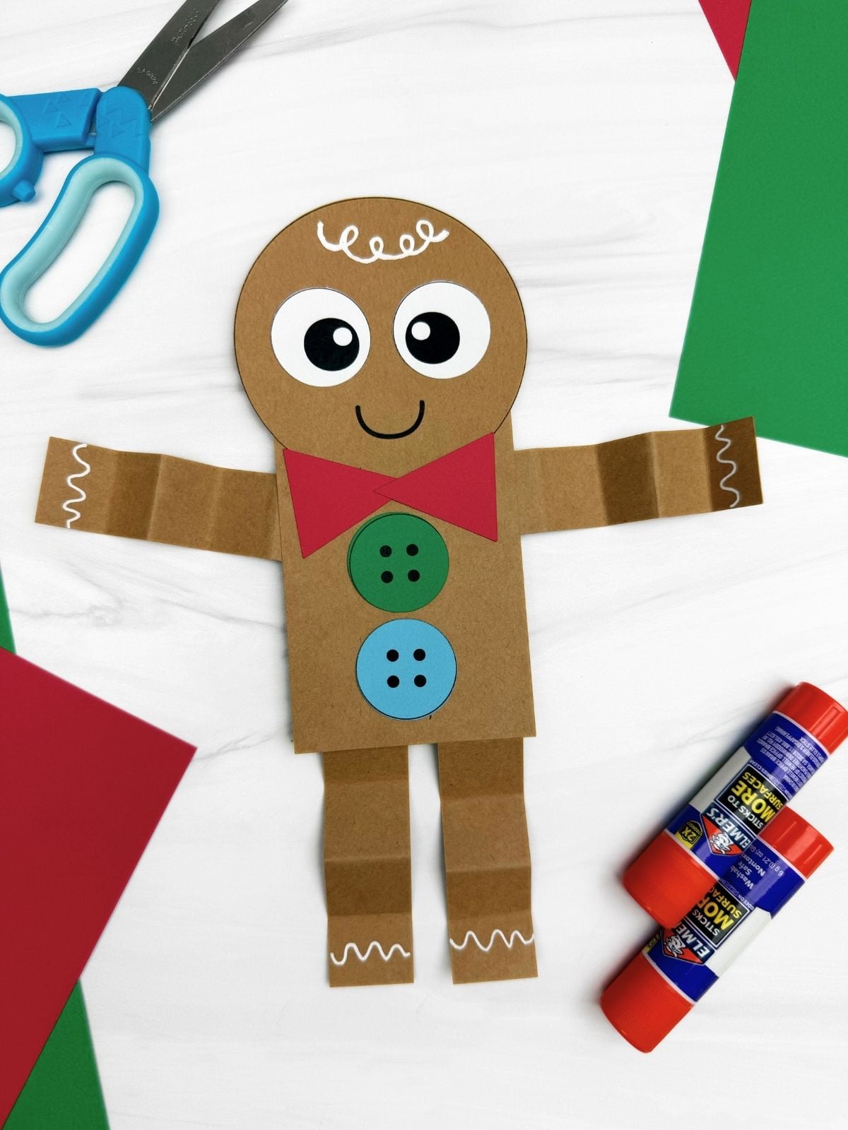 A completed Craft gingerbread man shape craft made from brown paper with googly eyes, red bowtie, colorful buttons, and folded arms and legs displayed with scissors, glue sticks, and colored paper for a Christmas kids craft.