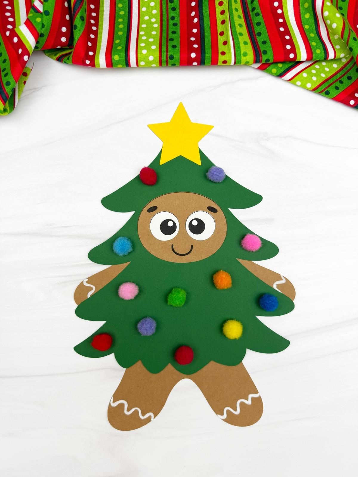 A completed Craft of a Christmas tree gingerbread disguise decorated with colorful pom poms, a smiling gingerbread face, green paper tree, and a yellow star topper for a fun kids Christmas craft.