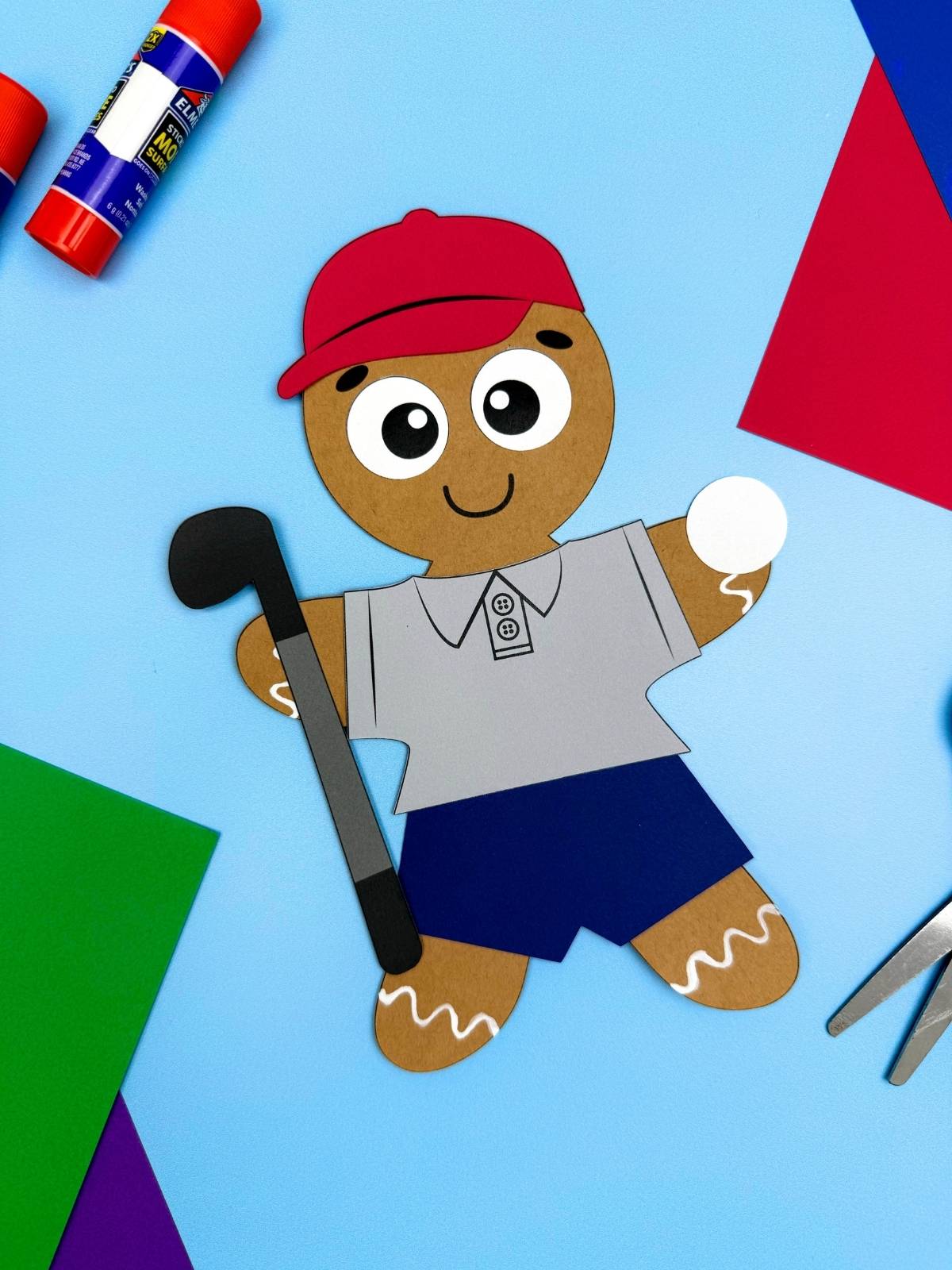 A completed craft showing a golfer gingerbread man paper craft with a red hat, gray golf shirt, blue shorts, golf club, and golf ball on a blue background with glue sticks, scissors, and colorful paper for a kids activity.