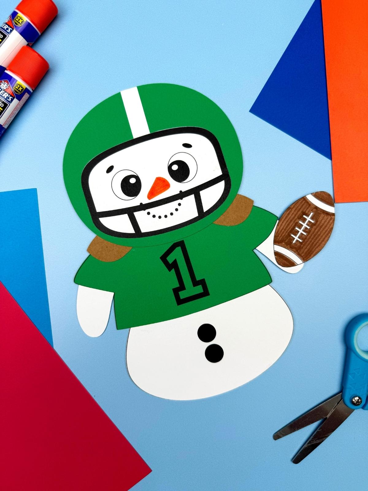 A completed Craft showing a football player snowman disguise with a green jersey and helmet, holding a paper football and surrounded by craft supplies for a Hide the Snowman kids