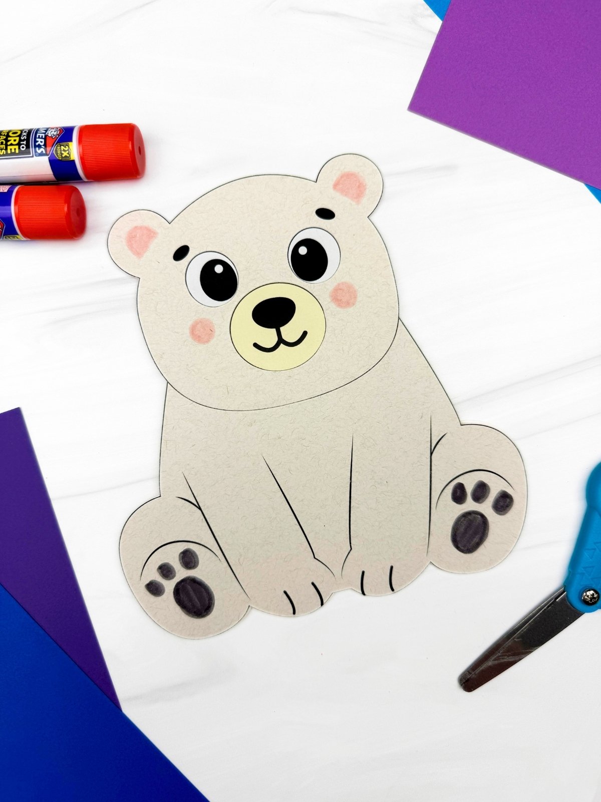 A completed craft showing a polar bear card made from a printable template displayed with scissors, glue sticks, and colored paper for a winter kids craft.