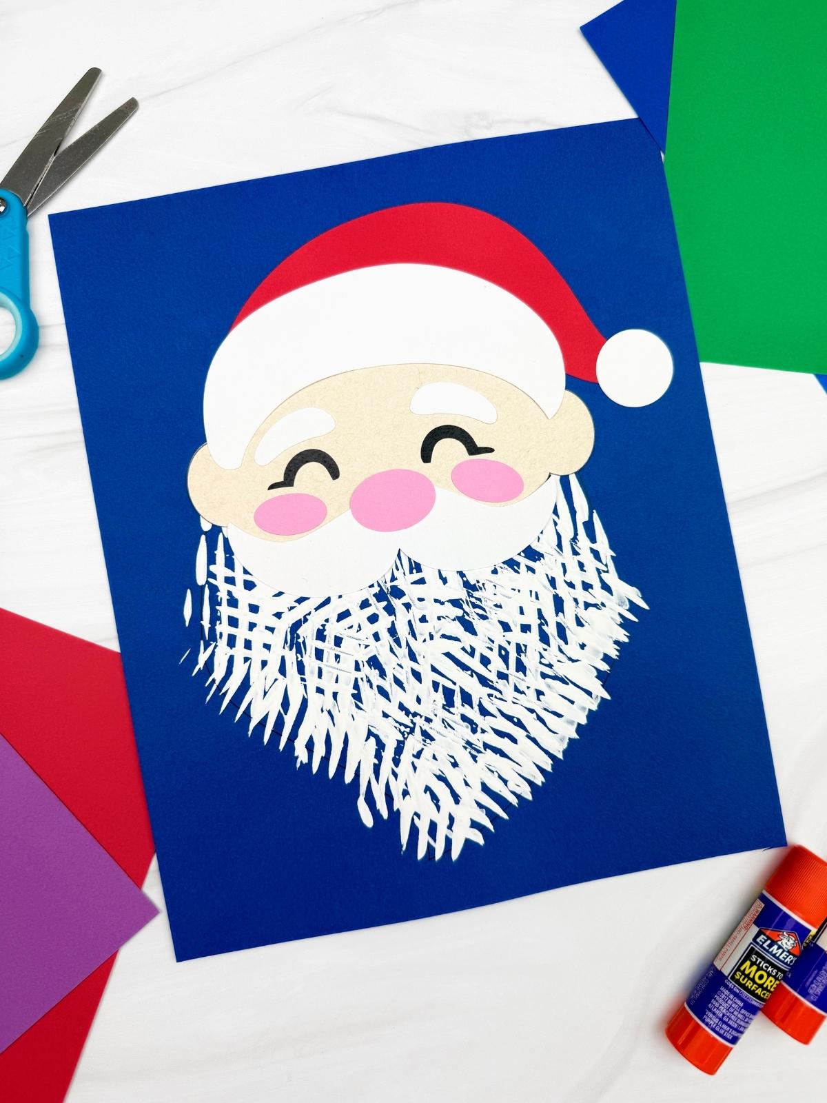 A completed Craft showing a fork painted Santa with a textured white beard, red hat, and pink cheeks on blue paper, surrounded by scissors, glue stick, and colorful cardstock for an easy kids Christmas Santa fork painting craft.