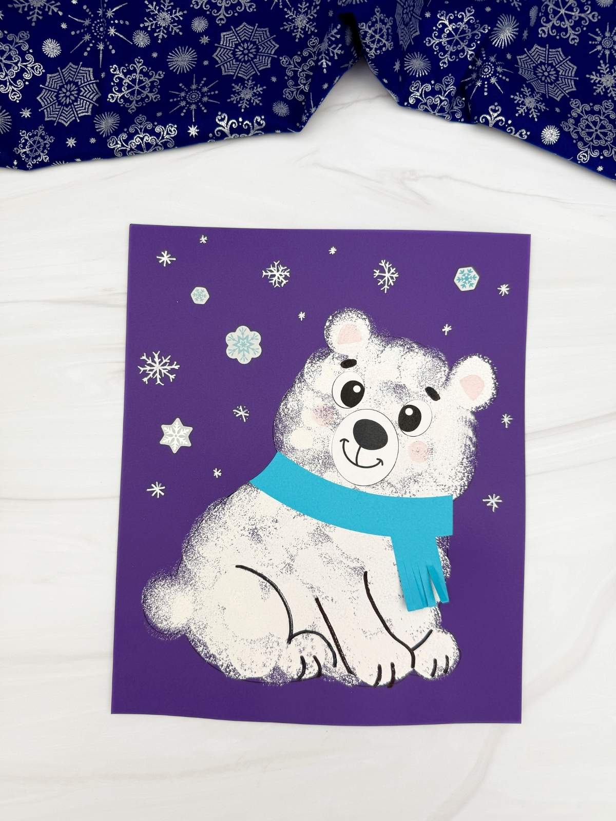 A completed craft featuring a sponge painted polar bear with a blue scarf on purple paper decorated with falling snowflakes for a winter kids craft.