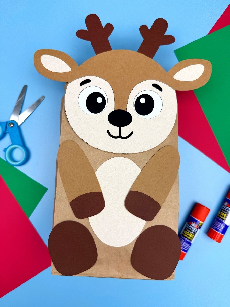 A completed stuffed paper bag reindeer craft displayed on a blue background with scissors, glue sticks, and colored paper, featuring a cute reindeer face, antlers, and paper legs.