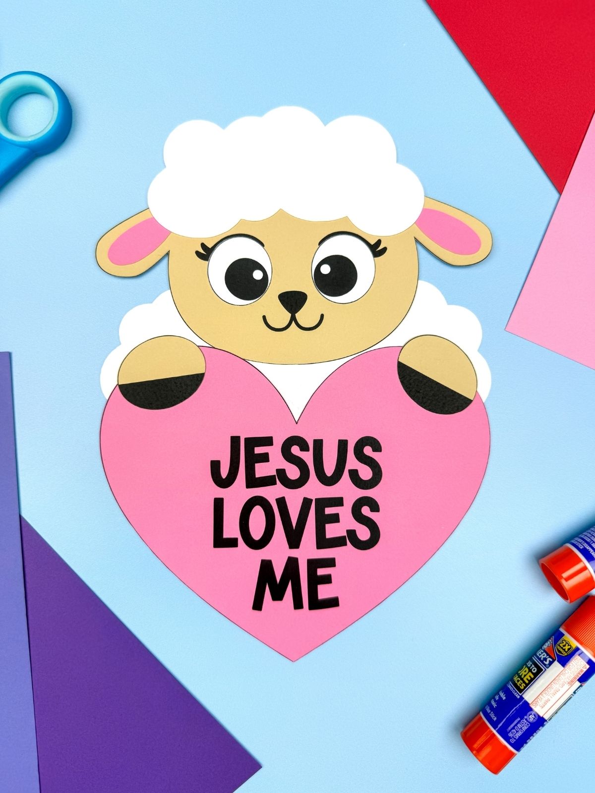A completed craft showing a Jesus Loves Me Valentine lamb holding a pink heart, easy Christian Valentine craft for kids displayed with paper and glue sticks on a blue background.