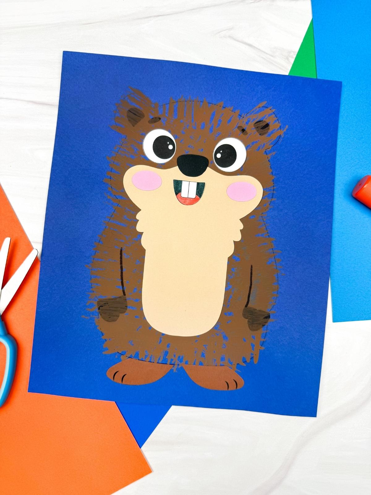 A completed craft showing a Groundhog Fork Painting with a cute brown groundhog on blue construction paper surrounded by colorful paper, scissors, and glue supplies.