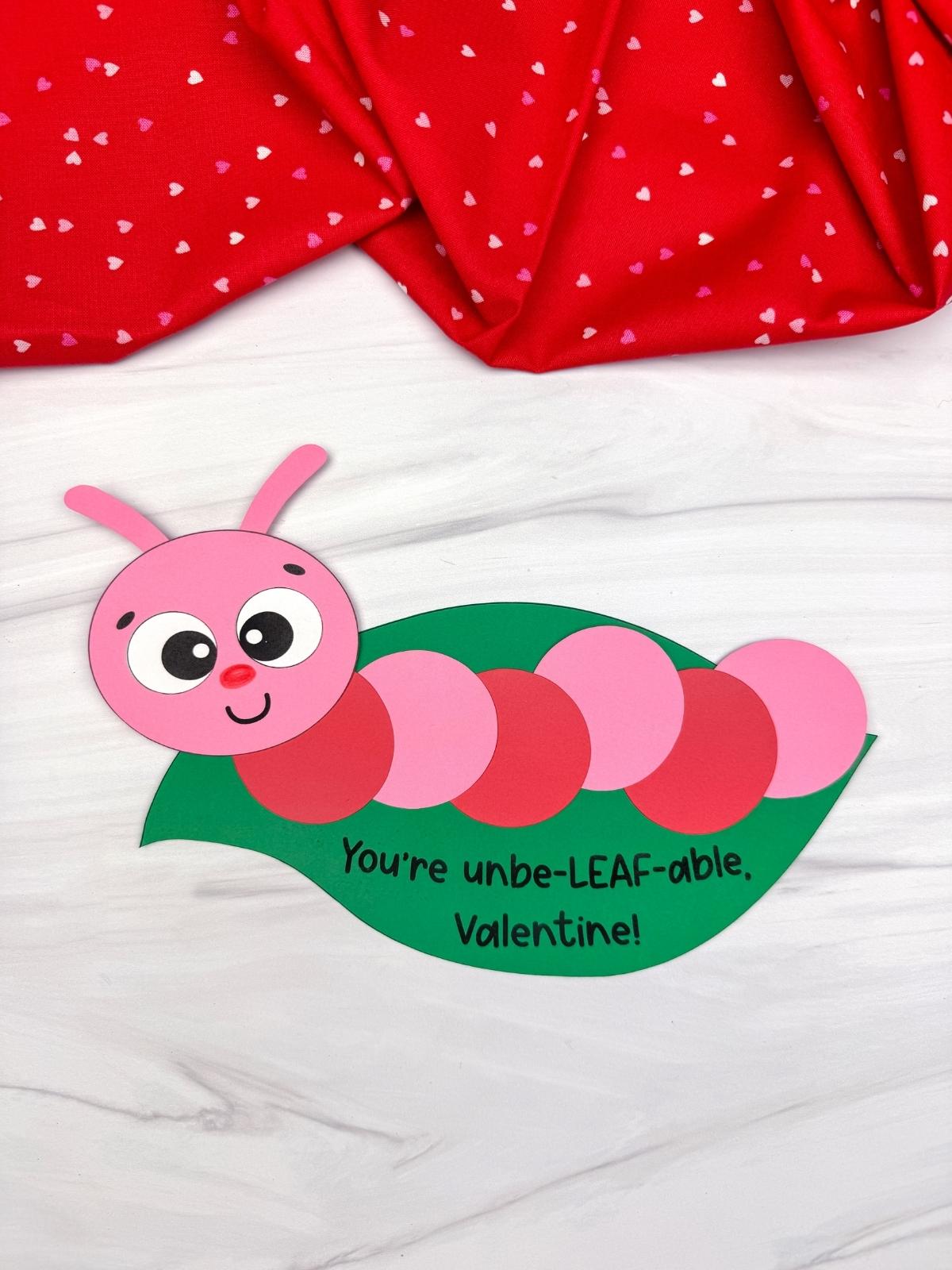A completed craft showing a Valentine caterpillar made from pink and red paper circles on a green leaf with a pun Valentine message, styled on a white background with red heart fabric for a kids Valentine caterpillar craft.