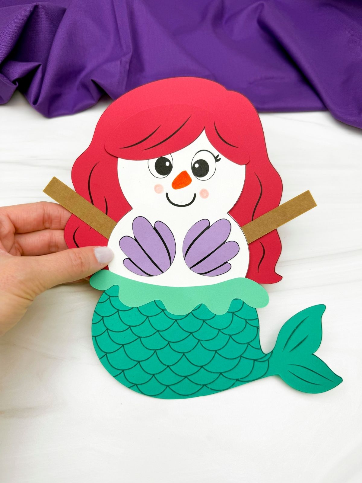 A completed craft showing a mermaid disguise a snowman paper craft with red hair, purple seashell top, green mermaid tail, and brown stick arms held up for a fun kids winter craft activity.