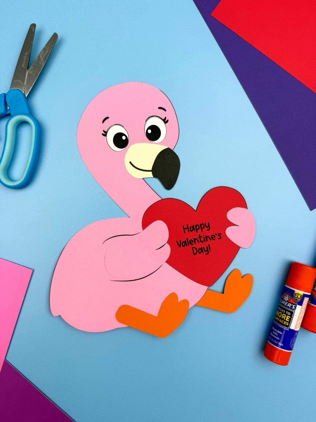 A completed Craft featuring a pink paper flamingo Valentine holding a red heart message on a blue background with scissors, glue stick, and colorful paper.