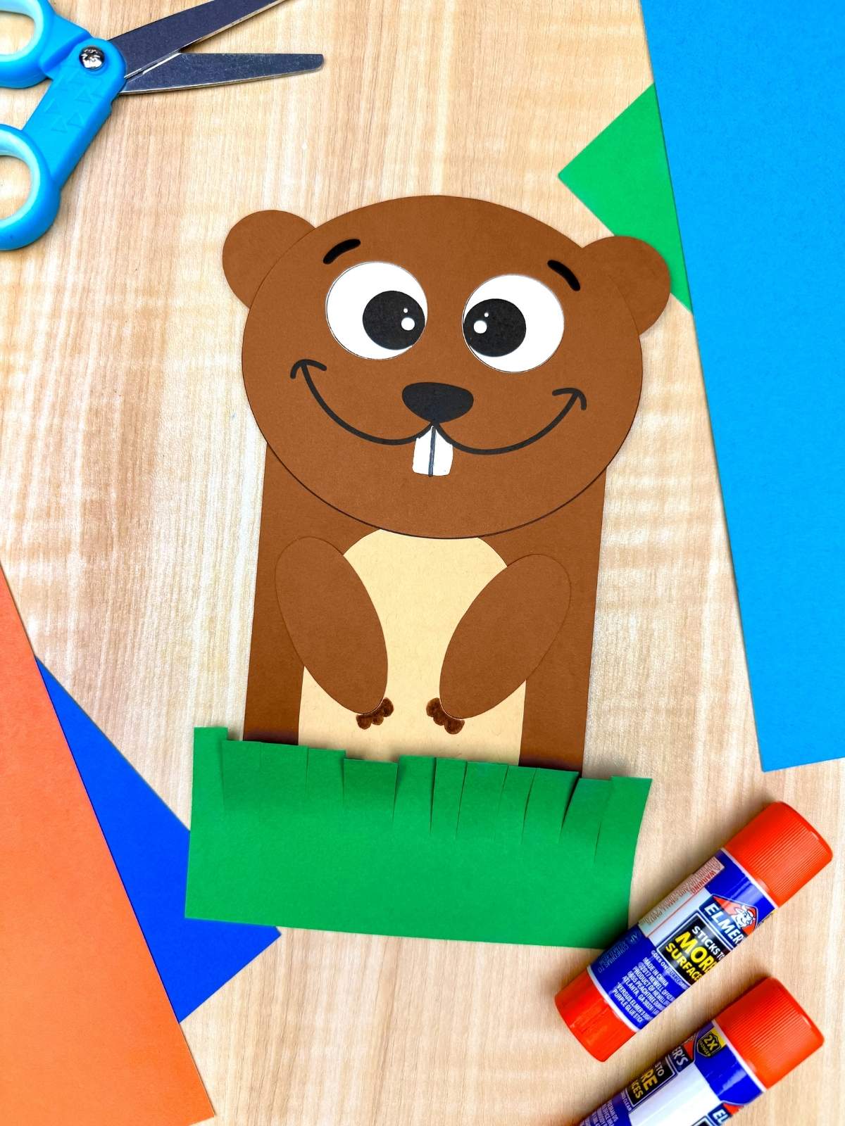 A completed Craft showing a smiling paper groundhog popping out of green grass with scissors, glue sticks, and colorful paper for an easy Groundhog Day craft for kids.