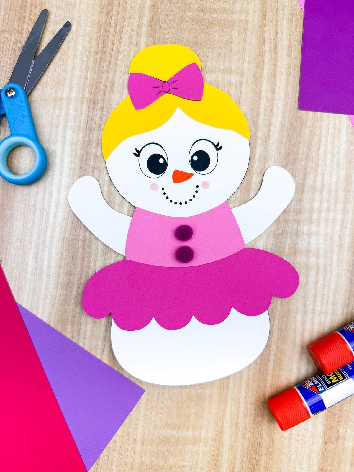 A completed craft showing a ballerina snowman disguise made from paper with a pink tutu, blonde hair with bow, pom pom buttons, and raised arms on a wooden table surrounded by scissors, glue sticks, and colorful cardstock.