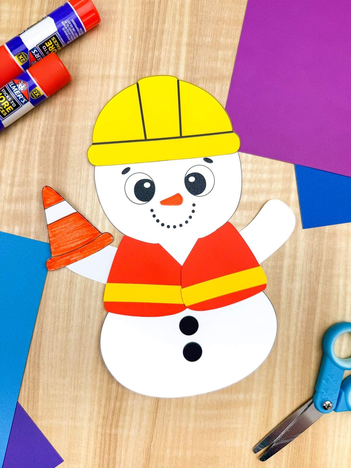 A completed craft showing a construction worker snowman made from paper, wearing a yellow hard hat, red safety vest, and holding an orange traffic cone, surrounded by glue sticks, scissors, and colorful cardstock for a kids winter craft activity.